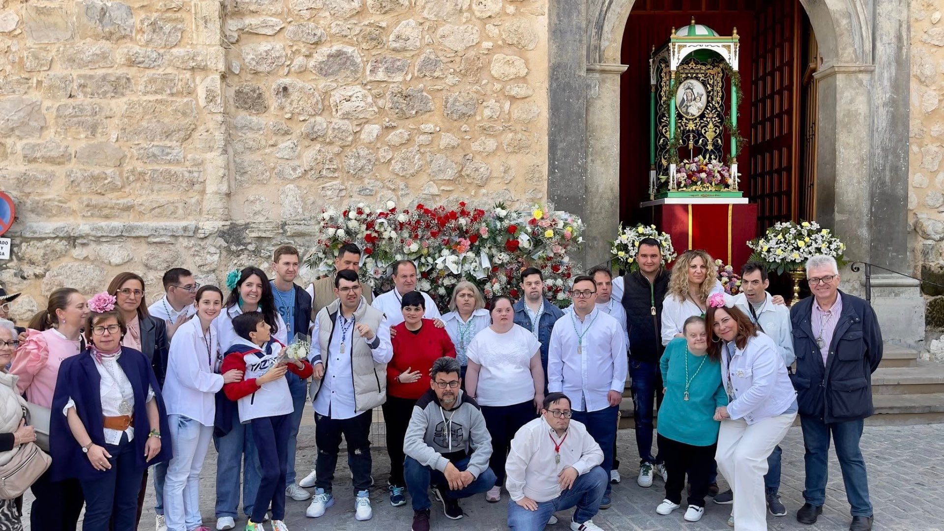 Fotografía real del grupo del Programa posando vestido de blanco junto a una gran ofrenda floral y la imagen de la Virgen, a las puertas de una iglesia de piedra de Torredelcampo.
