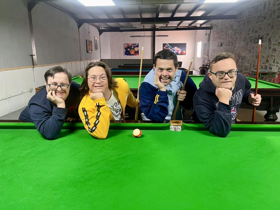 Fotografía real de cuatro personas del Programa apoyadas sobre una mesa de billar de tapete verde, sonriendo a cámara con tacos de billar y bola blanca en primer plano.