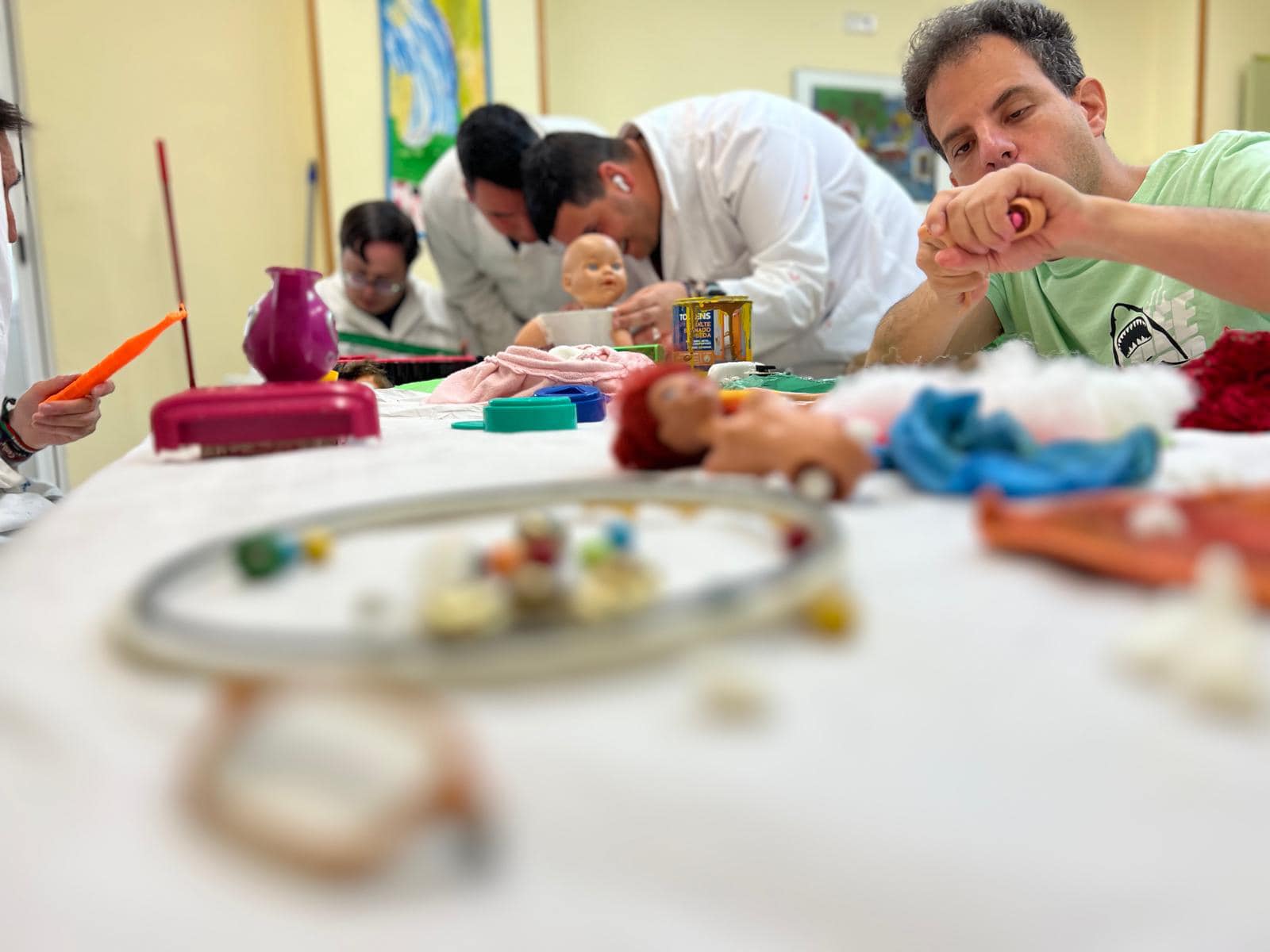Fotografía real de una mesa de taller con muñecos, peines, recipientes de colores y materiales sensoriales en primer plano, mientras participantes y profesionales con batas blancas trabajan al fondo.